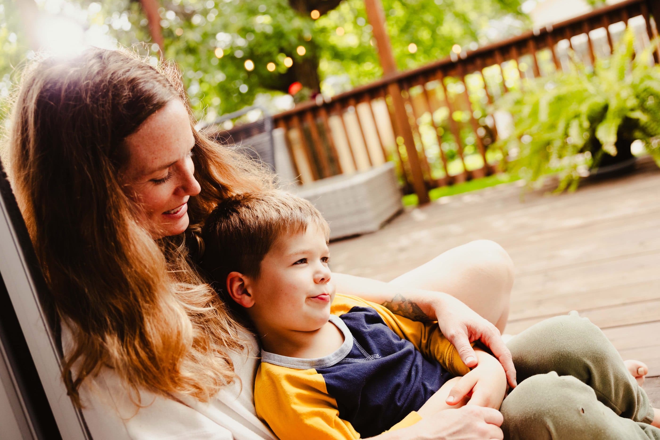 A mother and her young son sitting together on a sunny wooden deck