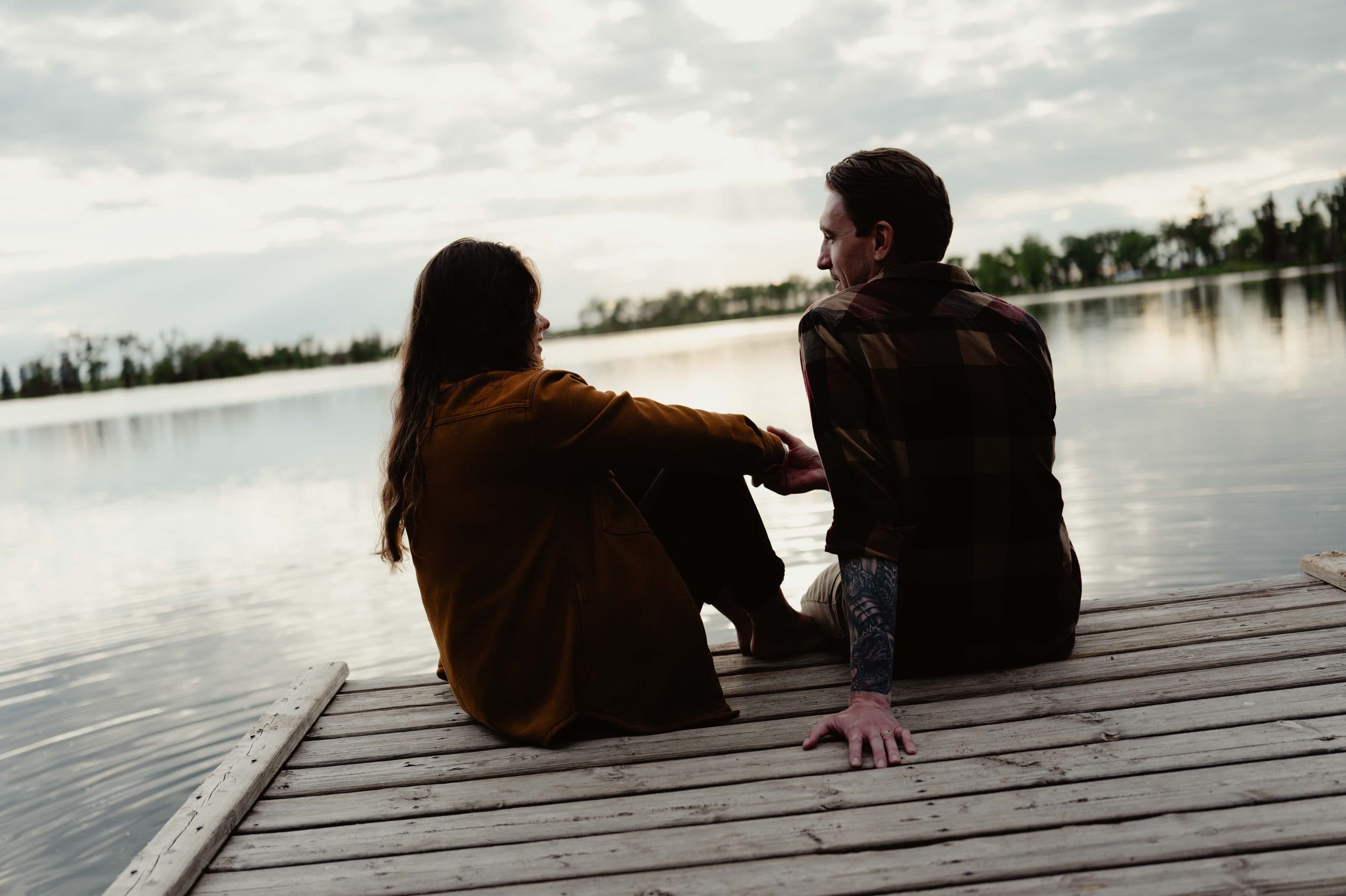 Two people sitting together at the end of a dock, looking out over a still lake