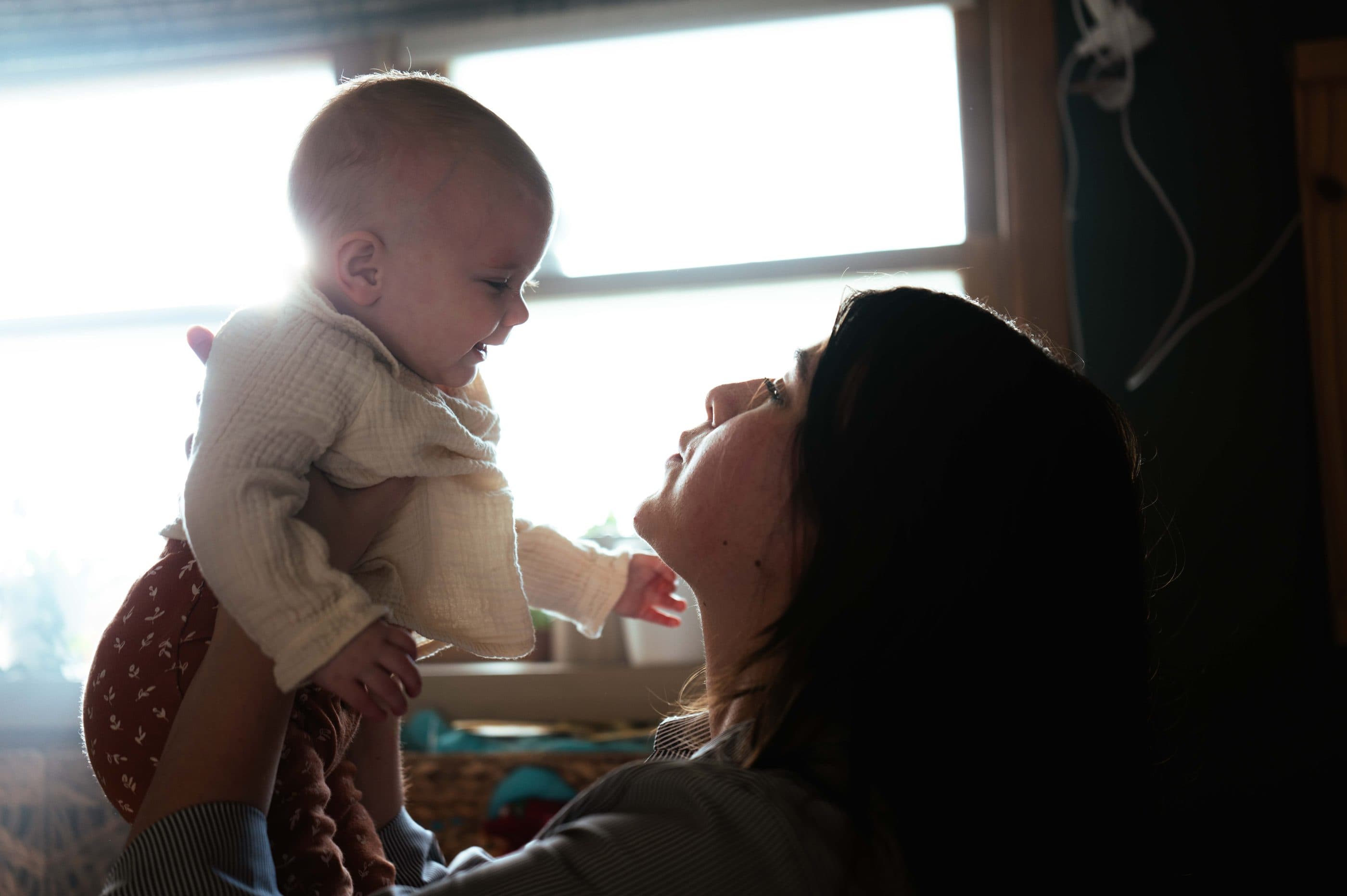 A mother lifting her baby toward the light of a window, both of them smiling