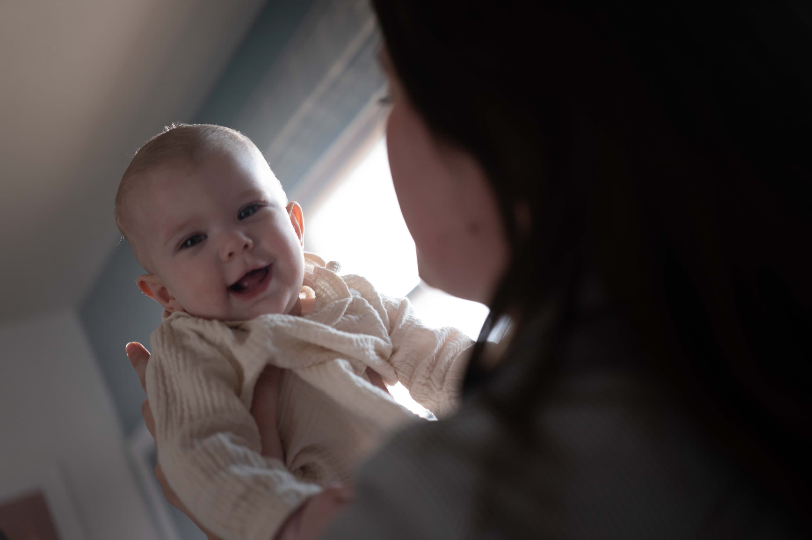 A mother lifting her smiling baby beside a sunny window