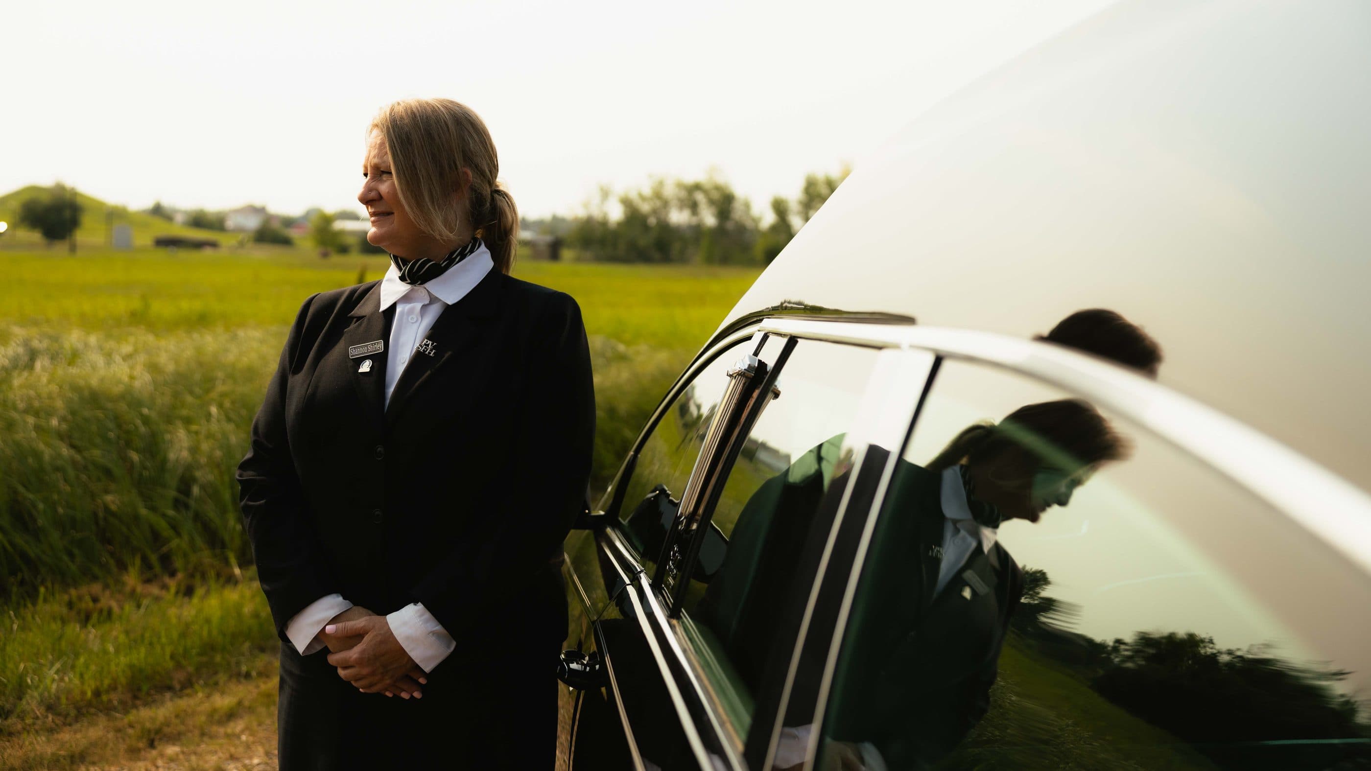 A funeral director beside the hearse on a prairie road at dusk