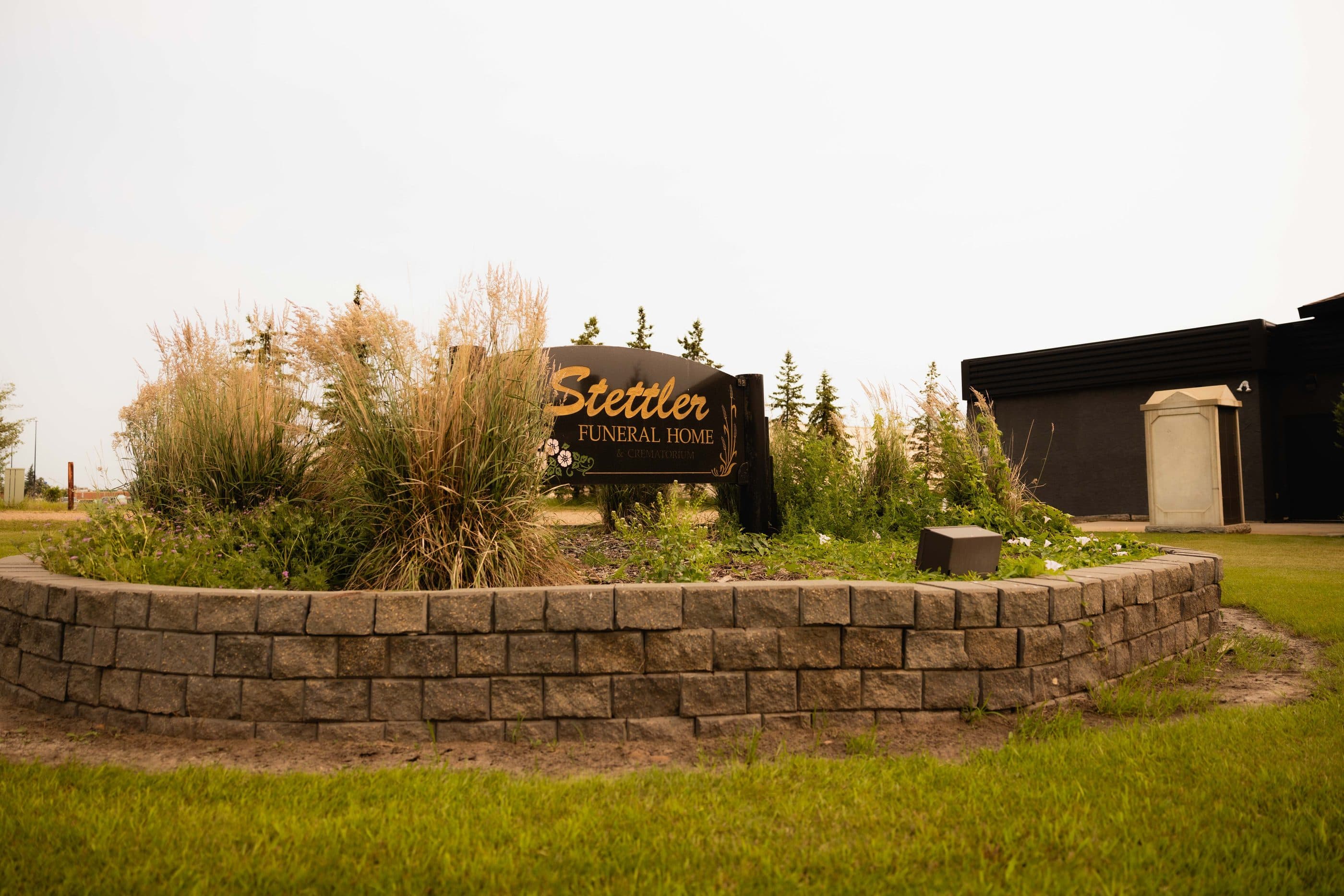The Stettler Funeral Home sign in its stone planter, with spruce along the drive