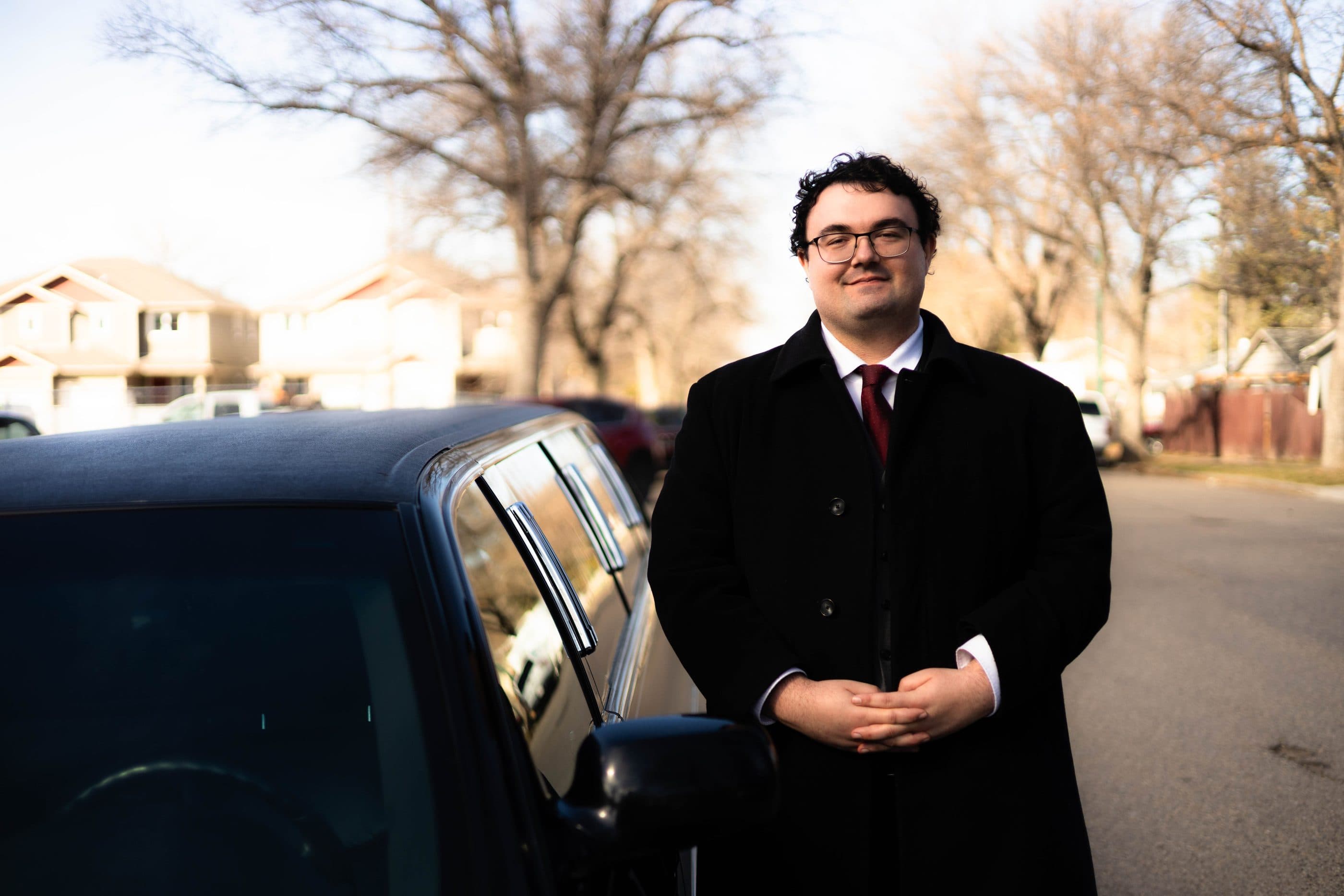 A funeral director standing beside the car on a tree-lined street